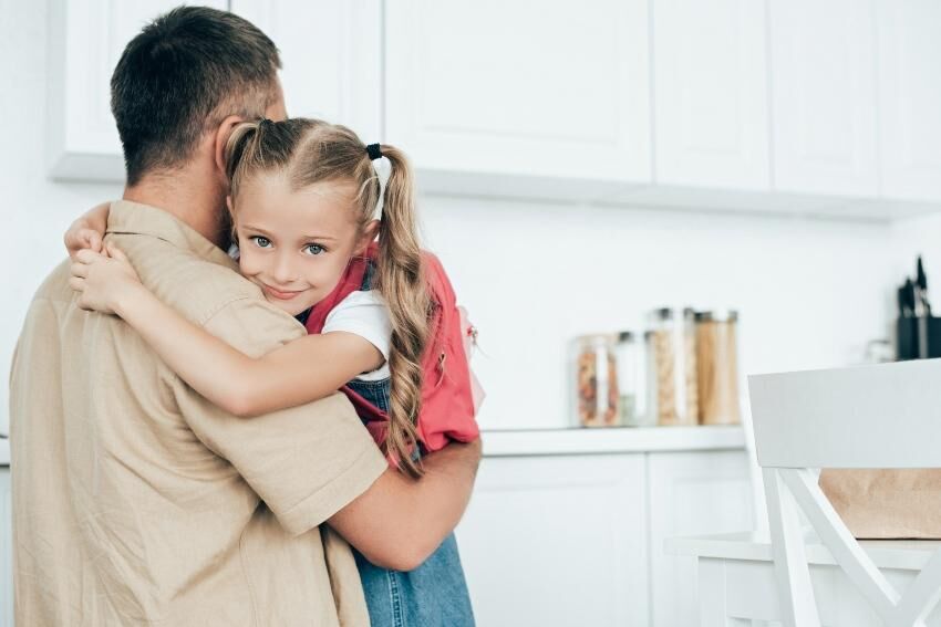 Für die emotionale Stabilität von Kindern ist Zuwendung das Wichtigste Vater umarmt seine Tochter, die einen Schulrucksack trägt